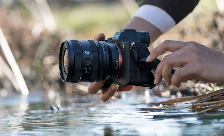 A usage image showing a user holding the α7 IV with the FE 16-25mm F2.8 G near the surface of a river. The user is shooting from a low position